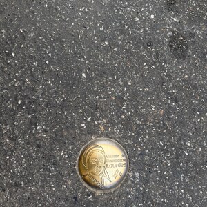 Puede incluir: Un marcador met&aacute;lico circular de color dorado incrustado en asfalto. El marcador presenta un perfil de una persona y el texto "Chemin de Bernadette Lourdes" en una fuente decorativa. Tambi&eacute;n hay dos peque&ntilde;as formas de estrella.