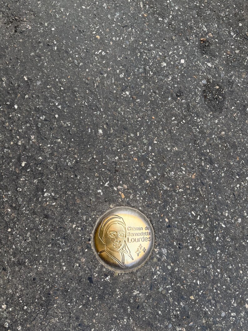 Puede incluir: Un marcador met&aacute;lico circular de color dorado incrustado en asfalto. El marcador presenta un perfil de una persona y el texto "Chemin de Bernadette Lourdes" en una fuente decorativa. Tambi&eacute;n hay dos peque&ntilde;as formas de estrella.