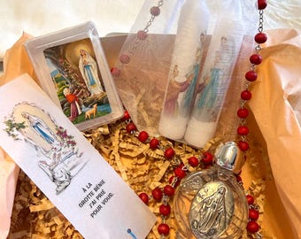 Puede incluir: Un set de regalo religioso que contiene un rosario con cuentas rojas, dos velas blancas en una bolsa de organza, una pequeña imagen enmarcada de la Virgen María y un marcapáginas de papel con el texto A LA GROTTE BENIE J'AI PRIE POUR VOUS.