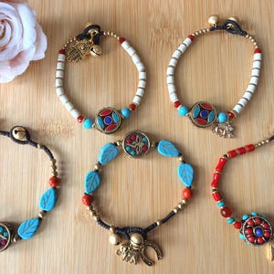 May include: An assortment of beaded bracelets with turquoise, red, and white beads. Some bracelets feature decorative charms and medallions with colorful accents. A pink rose is in the upper left corner, all on a wooden surface.