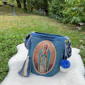 Puede incluir: Un bolso de hombro azul tejido a ganchillo con una imagen religiosa en la parte delantera. El bolso tiene un cierre de cord&oacute;n, una borla y un pomp&oacute;n. El bolso est&aacute; sobre una superficie blanca y esponjosa.