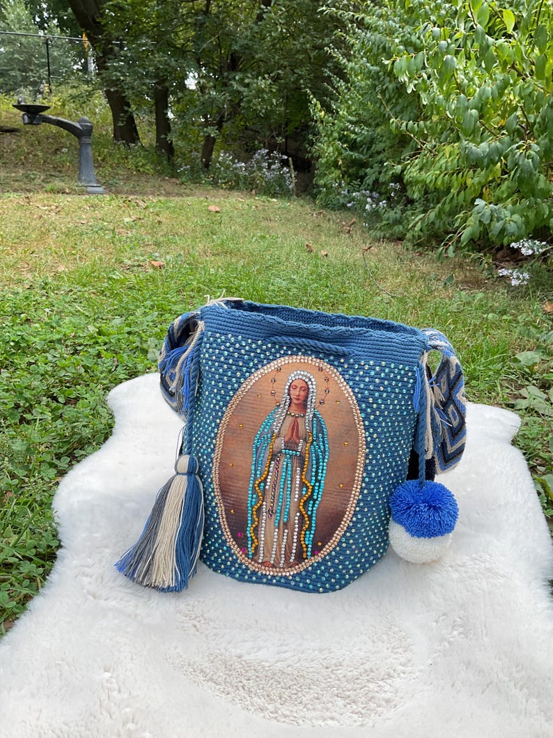 Puede incluir: Un bolso de hombro azul tejido a ganchillo con una imagen religiosa en la parte delantera. El bolso tiene un cierre de cord&oacute;n, una borla y un pomp&oacute;n. El bolso est&aacute; sobre una superficie blanca y esponjosa.