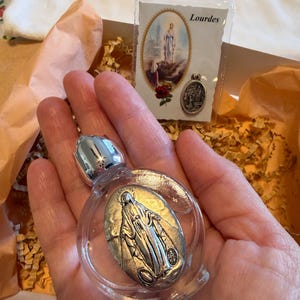 May include: A clear glass bottle with a silver cap and a silver oval medallion featuring a religious figure. A card with the word "Lourdes" and a small silver pendant are also visible.