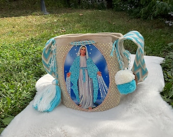 Puede incluir: Un bolso tipo cubo de ganchillo beige con una imagen ovalada central de la Virgen María rodeada de pequeños brillantes transparentes. El bolso tiene una correa ancha con un patrón de chevrón azul y blanco, acabado con borlas y pompones.