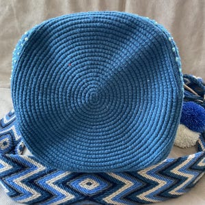 Puede incluir: Bolso azul tejido a mano con dise&ntilde;o circular. El bolso presenta una correa tejida con un patr&oacute;n geom&eacute;trico en tonos azules y blancos. Un pomp&oacute;n azul y un pomp&oacute;n blanco est&aacute;n unidos a la correa.