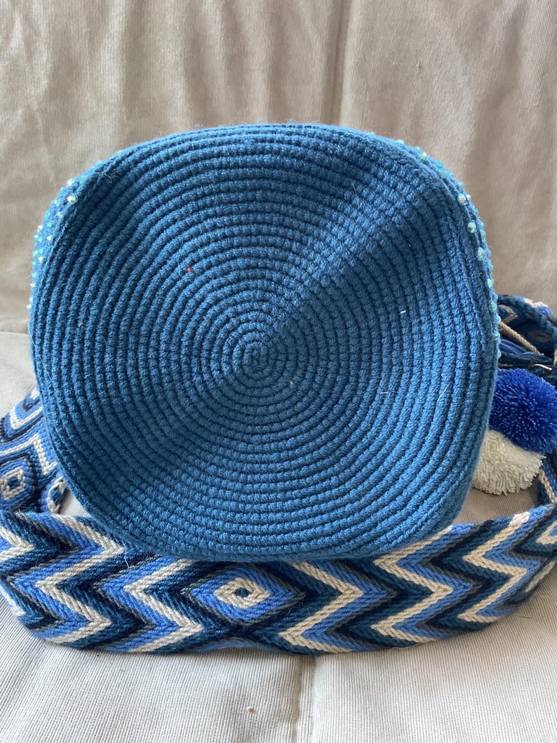 Puede incluir: Bolso azul tejido a mano con dise&ntilde;o circular. El bolso presenta una correa tejida con un patr&oacute;n geom&eacute;trico en tonos azules y blancos. Un pomp&oacute;n azul y un pomp&oacute;n blanco est&aacute;n unidos a la correa.