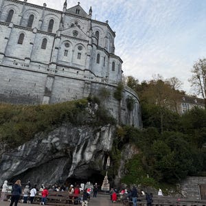 Puede incluir: Una gran iglesia de piedra gris con m&uacute;ltiples ventanas arqueadas y una cruz en la parte superior. Una entrada a una cueva es visible debajo, con gente reunida frente a ella. El cielo est&aacute; nublado.
