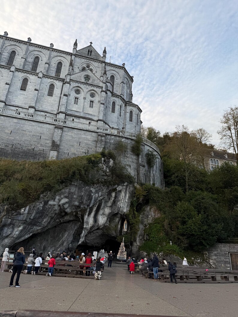 Puede incluir: Una gran iglesia de piedra gris con m&uacute;ltiples ventanas arqueadas y una cruz en la parte superior. Una entrada a una cueva es visible debajo, con gente reunida frente a ella. El cielo est&aacute; nublado.