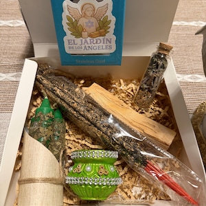 Könnte beinhalten: Eine Geschenkbox mit verschiedenen spirituellen Artikeln. Enthält eine türkisfarbene Box mit dem Text "El Jardin de Los Angeles", einen kleinen grünen Topf mit Perlen- und Goldakzenten, ein Glasfläschchen, Räucherstäbchen und eine Kerze.