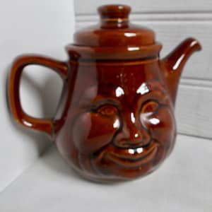 May include: A brown ceramic teapot with a smiling face. The teapot has a lid and a spout.