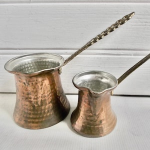 Op de afbeelding: Twee gehamerde koperen koffiepotten met lange handvatten. De grotere pot heeft een gedraaid handvat, terwijl de kleinere een recht handvat heeft.