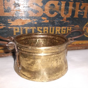 Puede incluir: Una pequeña olla de latón antigua con dos asas de hierro. La olla tiene un acabado martillado y está ligeramente deslustrada. La olla está sobre una superficie blanca.