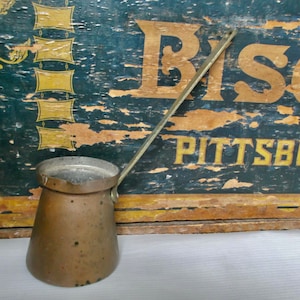 Op de afbeelding: Een vintage koperen koffiepot met een lange handgreep. De pot staat op een wit oppervlak voor een houten doos met de woorden "Biscuit Pittsburgh" erop geschilderd.