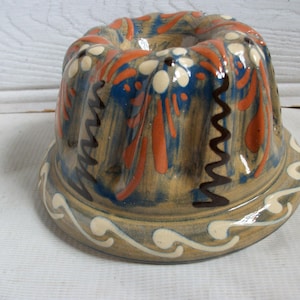 Puede incluir: Molde de cerámica para bizcocho con un diseño decorativo. La base es de color beige con patrones florales y abstractos azules, naranjas y blancos. Líneas en zigzag negras y un patrón de ondas blancas adornan el exterior.