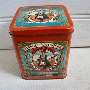 Puede incluir: Una lata cuadrada roja con una etiqueta azul y dorada. La etiqueta presenta a una mujer con un vestido azul y un delantal blanco, sosteniendo una cesta. El texto de la etiqueta dice "A La Belle Cantinière Mélange de Choix."
