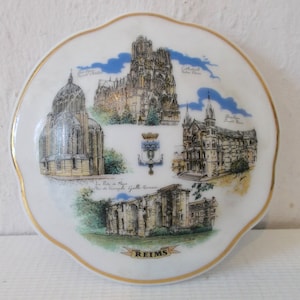 May include: A white ceramic plate with a gold rim features a colorful illustration of Reims, France. The plate depicts the Cathedral Notre Dame, the Basilica Saint Remi, the Porte de Mars, and the Gallo-Roman gate. The plate is inscribed with the word "REIMS" in gold lettering.