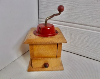 Puede incluir: Un molinillo de café vintage de madera con una parte superior de metal rojo y una manivela. El molinillo tiene un cajón para recoger el café molido.