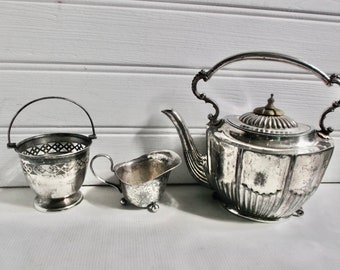 May include: A silver teapot, creamer, and sugar bowl with ornate designs. The teapot has a fluted body and a lid with a finial. The creamer and sugar bowl have a simple design with a rounded body and a handle.