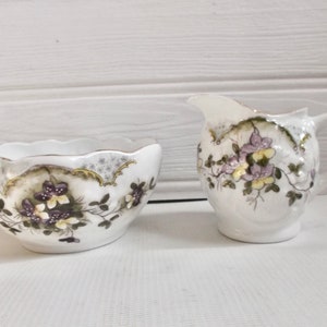 Puede incluir: Un juego de azucarero y jarra de crema de porcelana blanca con un diseño floral. El diseño presenta flores moradas, amarillas y verdes sobre un fondo blanco.