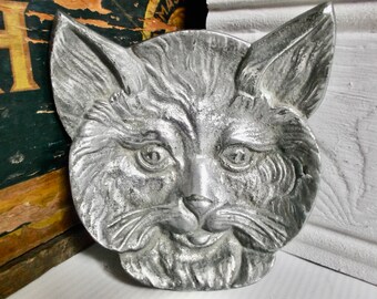 Puede incluir: Una decoración de pared de cabeza de gato de metal plateado con un diseño detallado y texturizado. Los ojos del gato son negros y la boca está ligeramente abierta.