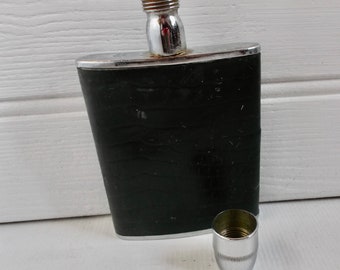 Puede incluir: Una petaca de plata y negra con tapón de rosca y tapón separado de plata. La petaca tiene un acabado negro texturizado.