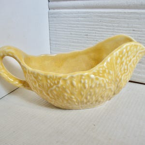 May include: A yellow ceramic gravy boat with a leafy design. The boat has a handle and a wide, shallow bowl.