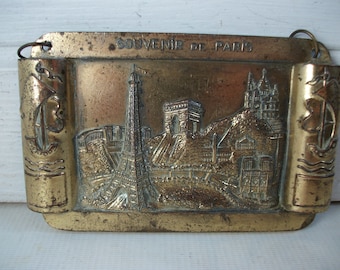 May include: A vintage brass souvenir plaque of Paris, featuring the Eiffel Tower, Arc de Triomphe, and Sacré-Cœur Basilica. The plaque has a rectangular shape with scroll-like side details and the words "SOUVENIR DE PARIS" at the top.