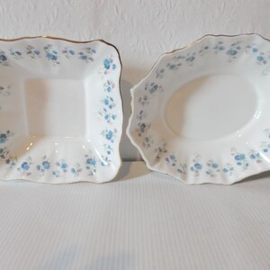 May include: Two white porcelain serving dishes. One is square, the other oval, both with a scalloped edge and a gold trim. Each dish is decorated with a pattern of small blue flowers and pink accents.