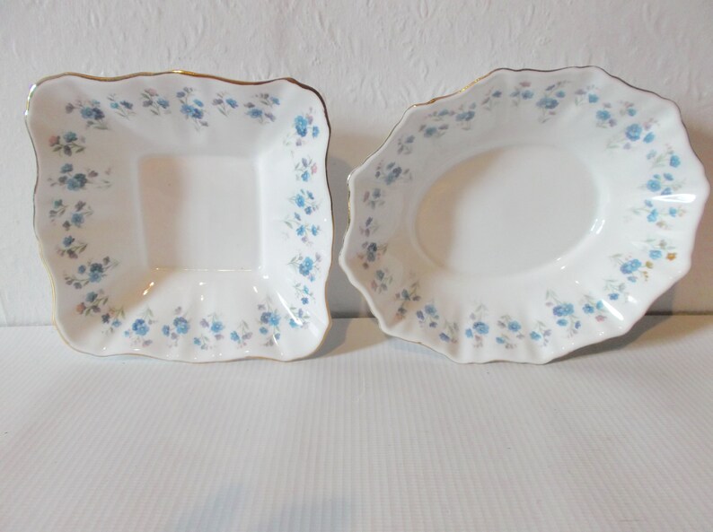 May include: Two white porcelain serving dishes. One is square, the other oval, both with a scalloped edge and a gold trim. Each dish is decorated with a pattern of small blue flowers and pink accents.