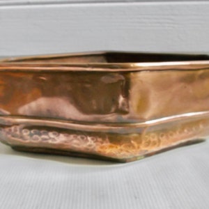 May include: A copper planter with a hammered finish. The planter has a square shape with rounded corners and a slightly flared lip.