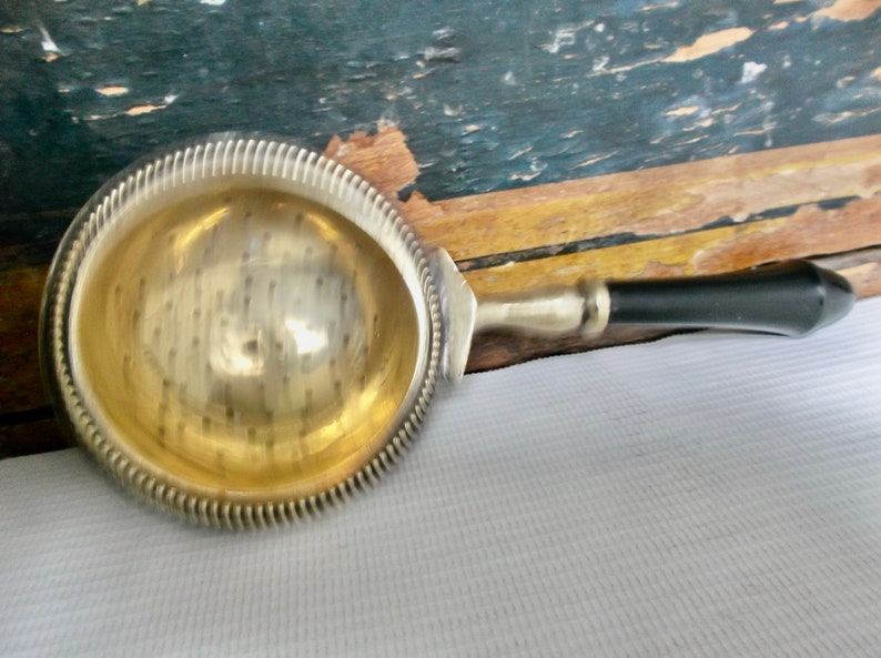 May include: A silver tea strainer with a black handle. The strainer has a round, perforated bowl with a decorative rim.