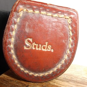 Puede incluir: Una funda de cuero marr&oacute;n con un borde cosido y la palabra "Studs" en relieve en letras doradas.