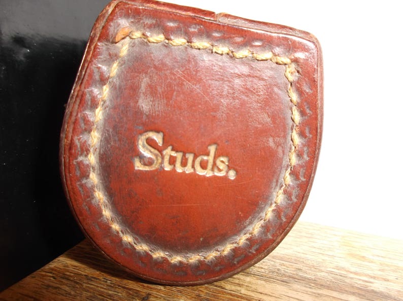 Puede incluir: Una funda de cuero marr&oacute;n con un borde cosido y la palabra "Studs" en relieve en letras doradas.