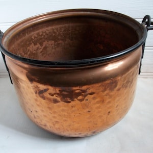 May include: A copper cauldron with a hammered texture and black metal handles. The pot has a wide opening and a rounded base. The interior is a darker shade of copper. The cauldron is likely used for cooking or decorative purposes.