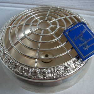 May include: Silver-plated Mayfair flower frog. The round bowl has a decorative rim and a wire grid top. A blue tag with gold lettering reads "Mayfair Silverplated Giftware."