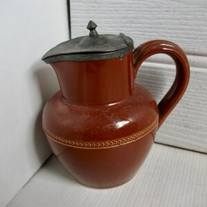 May include: A brown ceramic pitcher with a pewter lid. The pitcher has a decorative band around the middle and a handle.