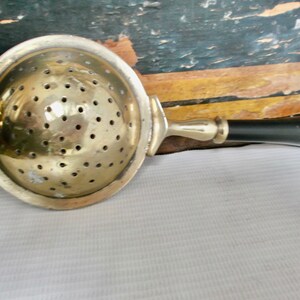 May include: A vintage silver tea strainer with a black handle. The strainer has a round, perforated bowl and a long handle.
