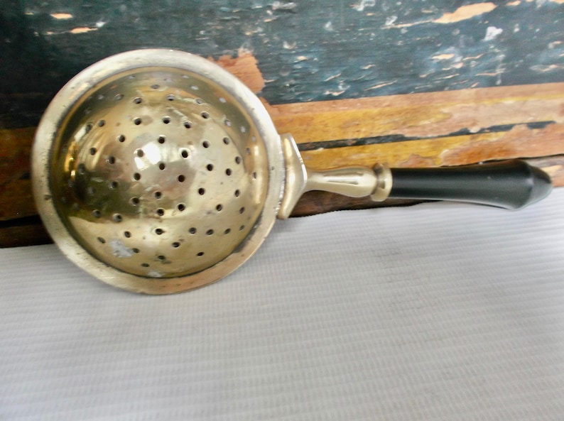 May include: A vintage silver tea strainer with a black handle. The strainer has a round, perforated bowl and a long handle.