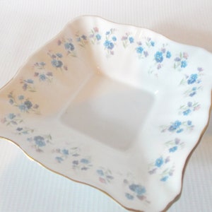 May include: A white, square-shaped dish with a scalloped edge and a gold trim. The dish is decorated with a delicate floral pattern of blue and purple flowers. A charming piece for serving or display.