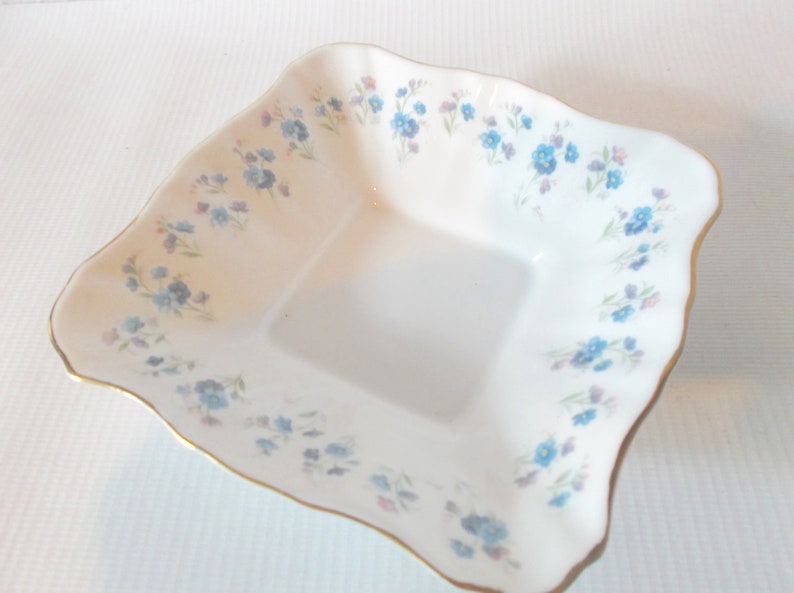 May include: A white, square-shaped dish with a scalloped edge and a gold trim. The dish is decorated with a delicate floral pattern of blue and purple flowers. A charming piece for serving or display.