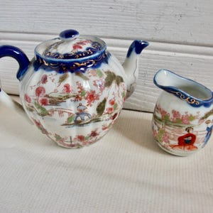 May include: A vintage porcelain tea set comprising a teapot and creamer. The teapot has a blue handle and lid with a floral design. The creamer has a matching design. The set is decorated with a landscape scene with red, pink, and green accents.