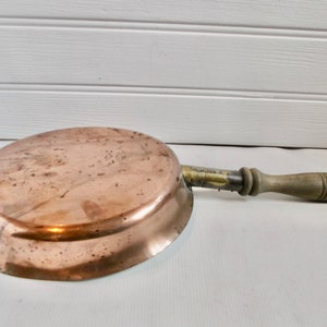 May include: A vintage copper frying pan with a wooden handle. The pan has a rounded shape and a slightly flared rim. The handle is attached to the pan with a metal band.