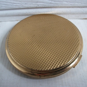 May include: A round, gold-toned compact with a textured, cross-hatched pattern on the lid. The compact has a shiny finish and a small clasp. The item is likely vintage and used for holding makeup.
