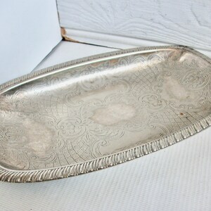 May include: An oblong silver-toned metal tray featuring an intricate engraved pattern of scrolling flourishes and cross-hatched lines. The tray has a raised, textured border with a repeating vertical ribbed design along the entire outer edge.