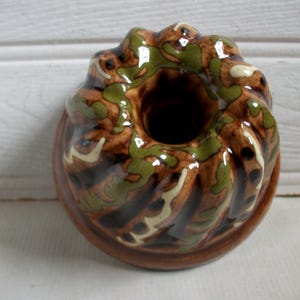 May include: A ceramic bundt cake mold with a brown base and a decorative top. The top features a pattern of green and white accents over a brown base. The mold has a central hole and a glossy finish.