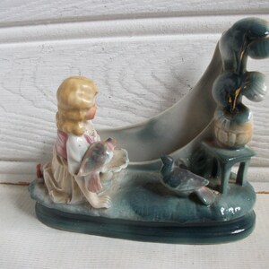 Op de afbeelding: Vintage keramiek beeldje met een knielend meisje met blond haar, dat een vogel vasthoudt. Het beeldje heeft een tealkleurige basis met een vogel en een decoratief element met een tafeltje en een plant. Het stuk is geschilderd in gedempte tinten.