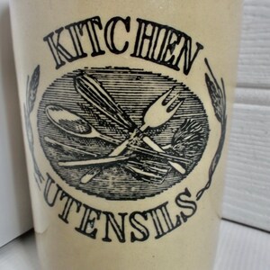 Pode incluir: Um pote de cerâmica branca para utensílios de cozinha com letras pretas que dizem "Kitchen Utensils". O pote tem uma ilustração em preto e branco de utensílios de cozinha, incluindo uma colher, um garfo, um batedor e uma espátula.
