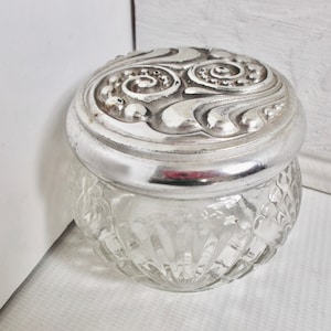 Könnte beinhalten: Eine klare Glasdose mit einem silbernen Deckel. Der Deckel hat ein dekoratives Design aus Spiralen und Punkten.