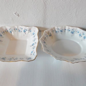 May include: Two white porcelain serving bowls. One is square, the other is oval. Both have a delicate blue floral pattern around the rim and gold trim. The bowls are empty and sit on a white surface.