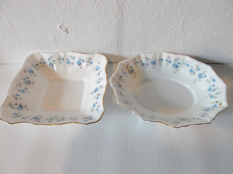 May include: Two white porcelain serving bowls. One is square, the other is oval. Both have a delicate blue floral pattern around the rim and gold trim. The bowls are empty and sit on a white surface.